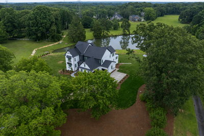 Aerial view of large white two-story home with dark roof on wooded lot beside a pond