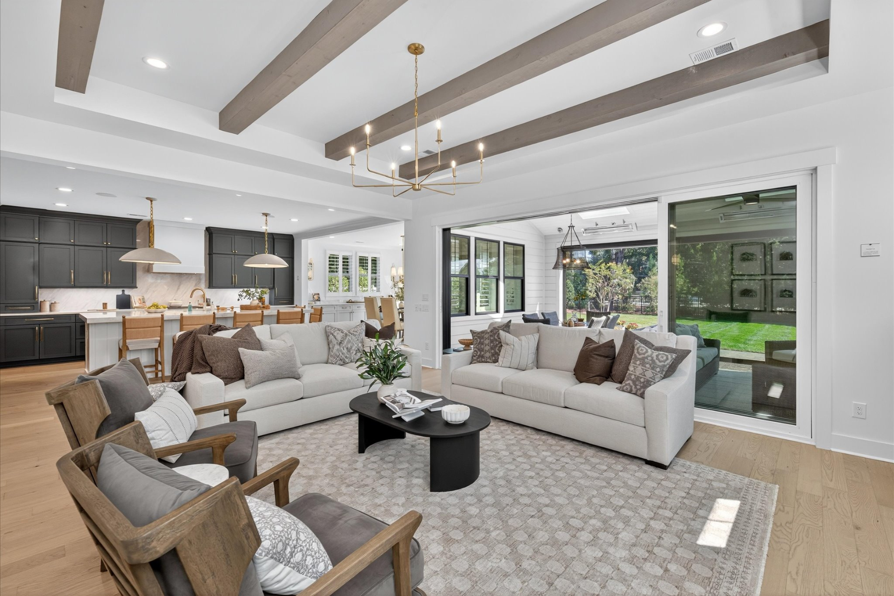 Open-concept living room with wood beam ceiling, light sofas, and view into kitchen and backyard