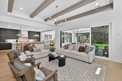 Open-concept living room with wood beam ceiling, light sofas, and view into kitchen and backyard