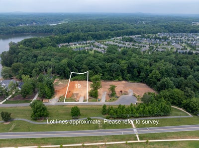 Aerial view of vacant lot 19 with cleared dirt, near a lake and residential neighborhood with wooded surroundings.