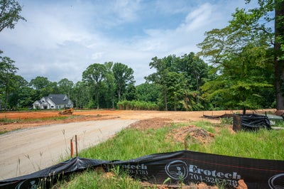 Cleared residential lot under construction with gravel road, excavator, and completed white home visible in background among trees.