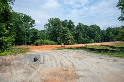 Cleared and graded residential lot with gravel driveway, red clay soil, and wooded tree line in background