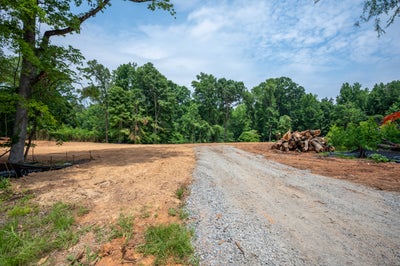 Cleared building lot with gravel driveway, cut logs stacked to the side, and wooded tree line in the background.