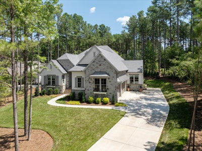 Single-story home with stone and stucco exterior, gray roof, two-car garage, curved driveway, and wooded surroundings.