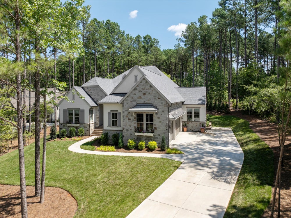 Single-story home with stone and stucco exterior, gray roof, two-car garage, curved driveway, and wooded surroundings.