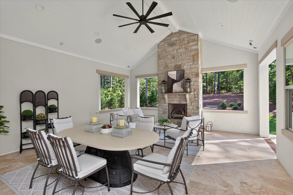 Covered patio with dining table, seating area, stone fireplace, vaulted ceiling, and ceiling fan overlooking wooded backyard