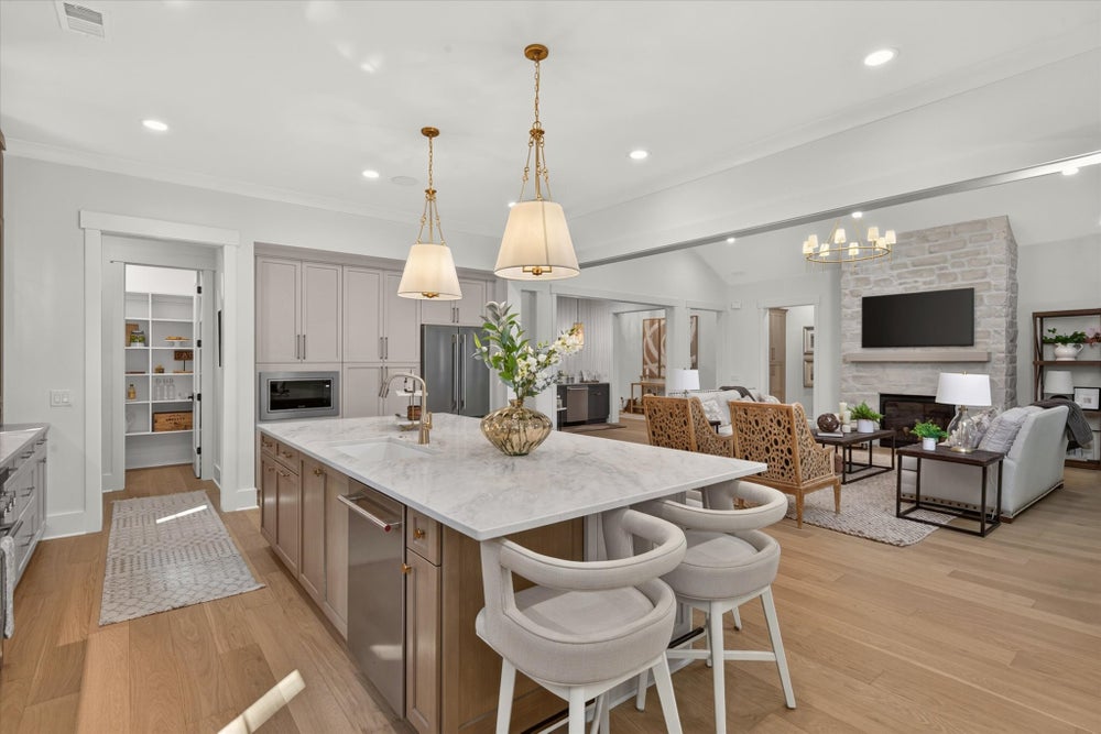Open-concept kitchen with large marble island, gray cabinets, and living room with stone fireplace in background