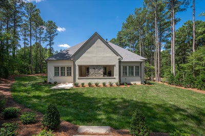 Single-story home with gray stucco exterior, covered porch with stone fireplace, and green lawn surrounded by tall pine trees.
