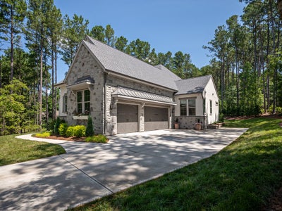 Stone and white exterior home with two-car garage, gray roof, and concrete driveway surrounded by trees