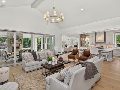 Open-concept living room with vaulted ceiling, gold chandelier, and sliding doors to outdoor patio, with kitchen visible in background