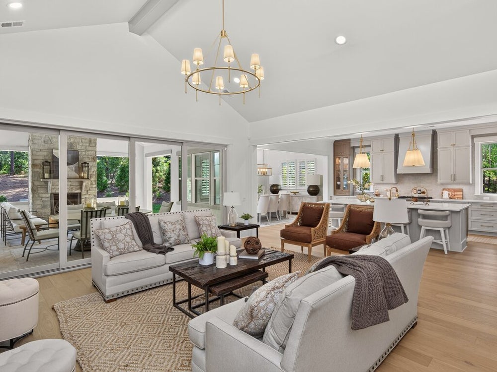 Open-concept living room with vaulted ceiling, gold chandelier, and sliding doors to outdoor patio, with kitchen visible in background