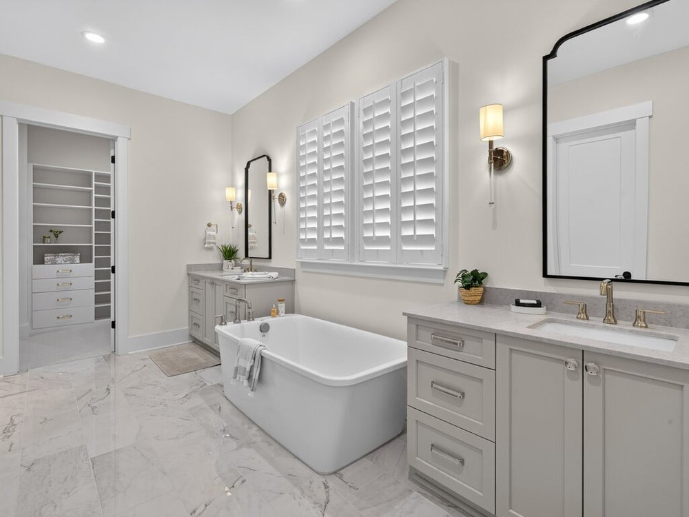 Master bathroom with freestanding soaking tub, dual vanities, marble tile floors, and plantation shutters with walk-in closet visible
