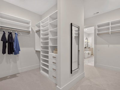Walk-in closet with white built-in shelving, hanging rods, drawers, and full-length mirror adjoining a bathroom