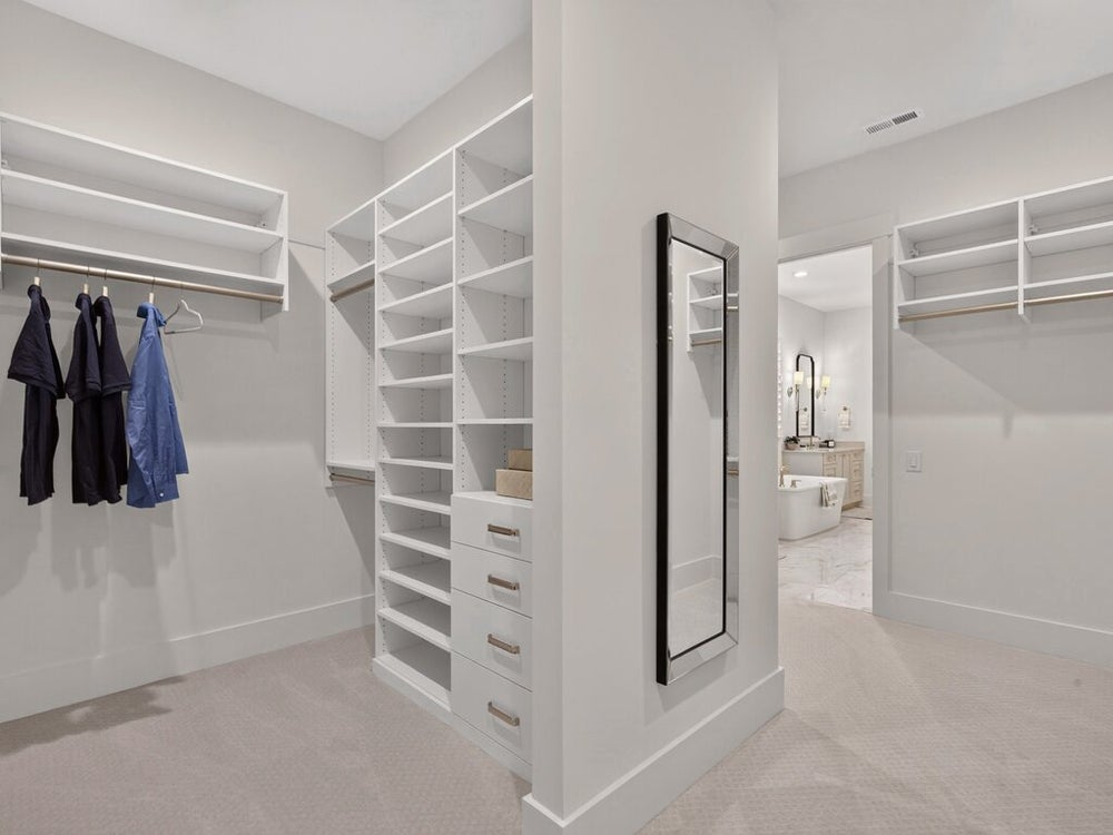 Walk-in closet with white built-in shelving, hanging rods, drawers, and full-length mirror adjoining a bathroom