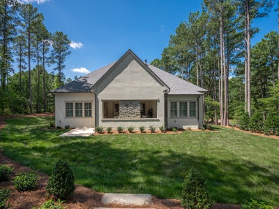 Rear exterior of single-story stucco home with stone fireplace, shuttered windows, patio, and wooded backyard