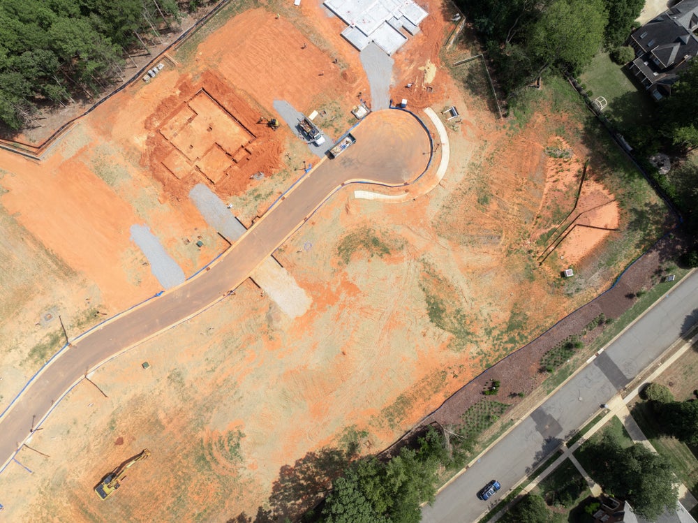 Aerial view of a residential construction site with graded red clay lots, paved road, and a home foundation in progress.