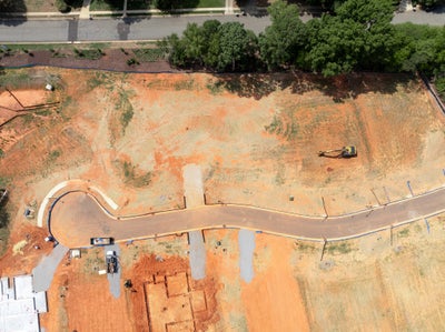 Aerial view of a residential construction site with cleared red clay lots, a curved road, and excavation equipment.