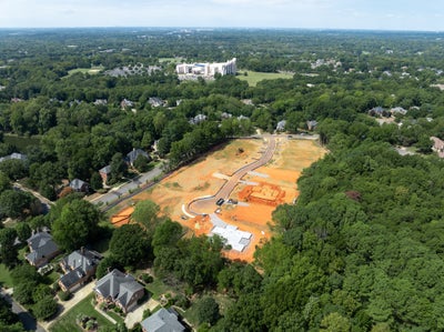 Aerial view of a residential development under construction with cleared lots, roads, and foundations surrounded by trees and existing homes.