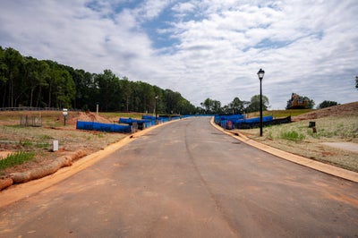 New subdivision road under construction with paved street, street lamps, blue erosion barriers, and graded lots beside a tree line.