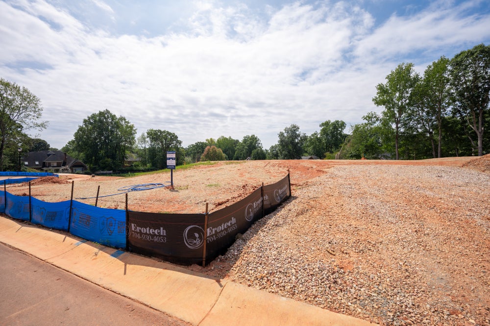 Cleared residential building lot with gravel, dirt, and erosion control fencing along the curb