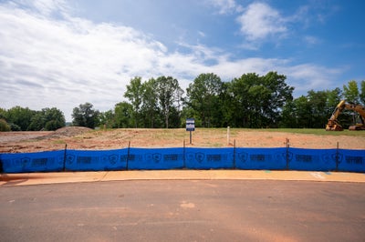 Empty residential lot with blue erosion control barrier, cleared dirt, and excavator visible at edge of tree line